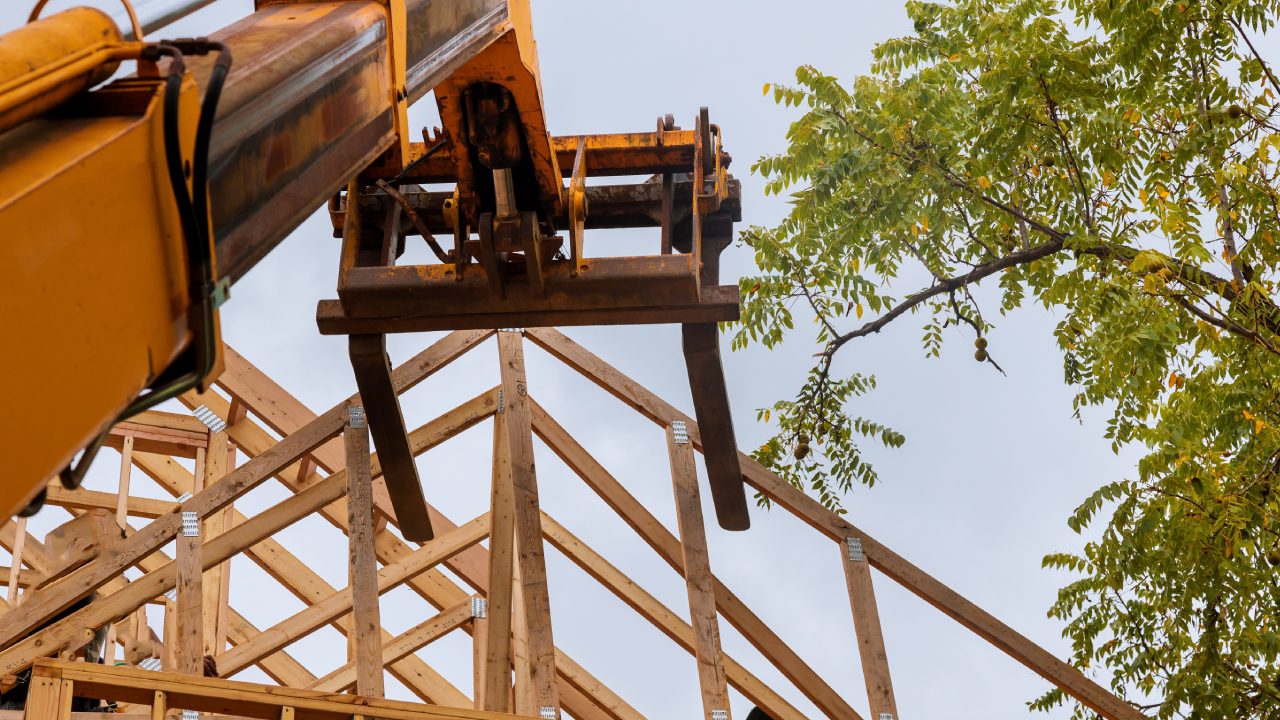 A construction crane lifting wooden beams to build a wooden roof structure against a clear sky.