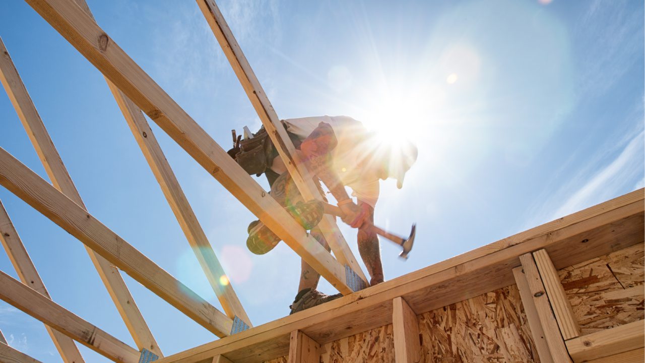 A construction worker using a hammer on a wooden frame under bright sunlight.